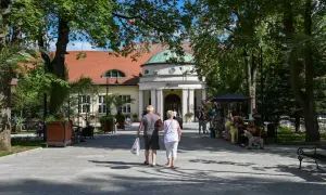 Para starszych osób spaceruje alejką przed budynkiem sanatorium.