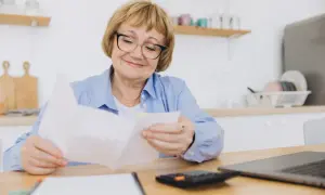 Starsza kobieta w okularach przegląda dokumenty przy biurku z laptopem i kalkulatorem.